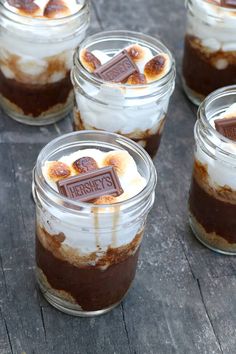 desserts in glass jars with marshmallows and chocolate on top, sitting on a wooden table