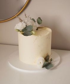 a white cake with flowers and greenery on top sits on a table next to a mirror