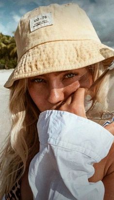 a woman wearing a hat on the beach with her hand to her face and looking at the camera