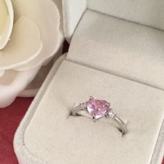 a pink heart shaped ring sitting in a box next to a white rose flower on a red surface