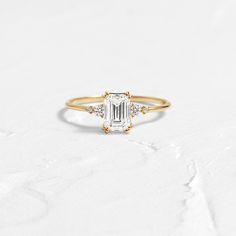 a gold ring with an emerald cut diamond and three small diamonds on the band, sitting on a white surface