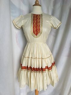 "Vintage Square Dancing/Patio Dress makes a fabulous addition to the wardrobe with unique red/gold embroidered bodice and gold metallic rick-rack trim of yesteryear! Pretty dress shows a rounded neckline w/ a festive embellished bodice and darts for better fit. The short sleeves are also trimmed w/ the gold rick-rack. Waistline has pretty fabric piping detail and is elasticized in back. Dress is gathered at the waist and three gathered tiers flow adorned w/ more of the red embroidered trim w/ go Festival Dresses With Embroidered Border And Fitted Stretch, Ceremonial Fitted Dress With Embroidered Border, Festive Fitted Dress With Embroidered Border, Fitted Bollywood Dress With Embroidered Border, Festive Embroidered Border Fitted Dress, Fitted Embroidered Dress For Ceremonial Festive Occasions, Festive Ceremonial Fitted Embroidered Dress, Fitted Bollywood Cream Dress, Fitted Cream Bollywood Dress