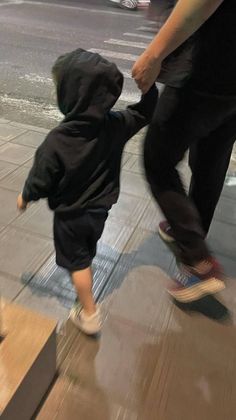 a small child in a black hoodie is walking down the sidewalk with his foot on a skateboard