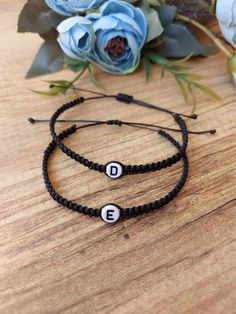 two black cord bracelets with white letters on them and blue flowers in the background