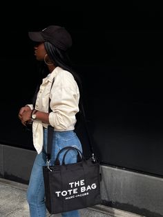 Black girl with Black Marc Jacobs The Tote Bag, wearing blue American Eagle high-waisted jeans, yellow light jean jacket, black adidas baseball cap and black Anne Klein watch Thetotebag Marc Jacobs, Marc Jacobs Tote Bag Small Vs Mini, The Tote Bag Outfit Marc Jacobs, The Tote Bag Marc Jacobs Outfit, Mark Jacobs Tote Bag, The Tote Bag Outfit, Black Marc Jacobs Tote Bag, Outfit With Tote Bag