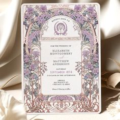 an ornate wedding card with purple flowers and vines on the front, is displayed next to some feathers