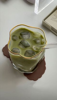 a green liquid in a glass with ice cubes on the rim next to a book