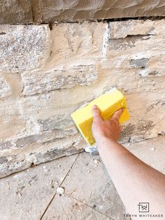 a hand holding a yellow sponge on top of a brick wall next to a cement block