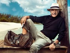 an older man sitting on a wooden bench next to the ocean with a bag in front of him