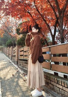 a woman standing next to a brick wall in front of trees with orange leaves on it