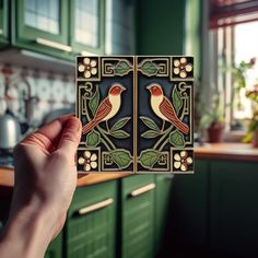 a hand holding up a decorative tile with two birds on it in a kitchen area