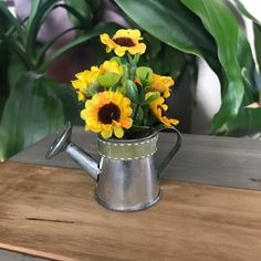 a metal watering can filled with yellow flowers on top of a wooden table next to potted plants