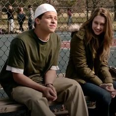 a man and woman sitting on a bench in front of a tennis court while one looks at the camera