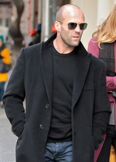 a bald man wearing sunglasses and a black coat is walking down the street with his hands in his pockets