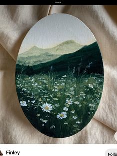 an oval painting with white flowers in the foreground and green mountains in the background