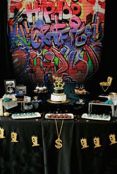 a table topped with cakes and desserts next to a wall covered in graffitti