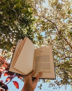 a person is holding an open book in front of some trees with leaves on it