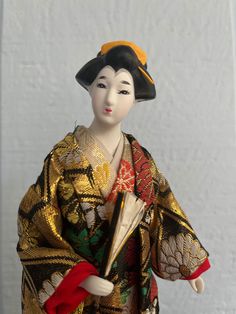 This vintage Japanese doll is dressed in traditional Japanese silk attire, specifically a kimono with an intricate pattern featuring gold, black, red, and other multi-colors, which gives this doll girl royal temperament. The kimono has wide sleeves and is accessorized with a red obi (sash) tied at the back.  The gown has so many layers while so neat on the doll. Amazing! The doll is also wearing a headpiece that matches the kimono's pattern. The doll girl's face is carefully hand-painted to life Traditional Gold Ceremonial Kimono, Obi Sash, Jensen Beach, Japanese Doll, Kimono Pattern, Japanese Silk, Japanese Dolls, Traditional Japanese, Wide Sleeves