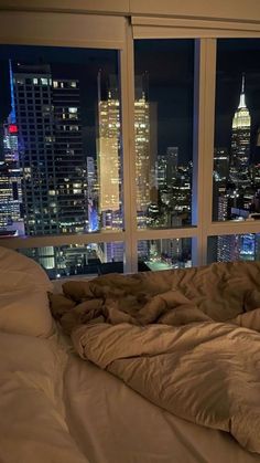 an unmade bed in front of a large window overlooking the city skyline at night