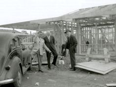 two men standing next to an old car in front of a building under construction with another man looking on