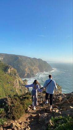 two people walking up a rocky path near the ocean on a sunny day with mountains in the background