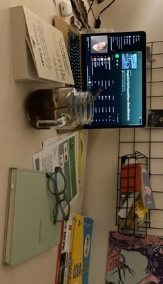 a laptop computer sitting on top of a desk next to books and glasses with eyeglasses hanging from the wall