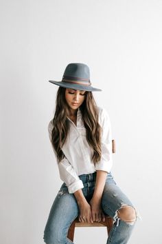 a woman sitting on top of a wooden chair wearing a hat and ripped denims