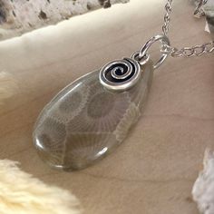 Embrace the natural beauty of Michigan with this Handcrafted Petoskey Stone Cabochon Pendant. This stunning piece features a beautifully polished Petoskey stone, skillfully shaped into a cabochon and adorned with a drilled hole for versatility. Attached to a durable stainless steel jump ring, this pendant is embellished with a charming swirl charm, adding a touch of whimsy and elegance to the design. Measuring approximately 0.75 x 1.2 inches, each stone is uniquely crafted, showcasing the distin Nickel-free Sterling Silver Swirl Necklace, Handmade Stainless Steel Round Pendant Charm Necklace, Adjustable Spiral Necklace Nickel Free, Silver Swirl Necklace For Gifting, Adjustable Spiral Nickel-free Necklace, Silver Swirl Necklace For Gift, Silver Swirl Necklace Perfect For Gifts, Unique Swirl Jewelry As A Gift, Handmade Swirl Necklace For Gift