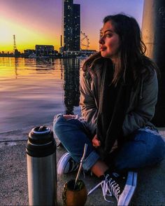 a woman sitting on the ground next to a water fountain at sunset with buildings in the background