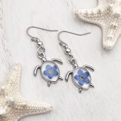 Transport yourself to the tranquil shores with our enchanting Forget Me Not Sea Turtle Earrings, blending playful charm with coastal elegance. Crafted with exquisite detail, they capture the essence of sun-kissed beaches and azure waves. Whether lounging by the shore or exploring coastal treasures, these earrings are a delightful keepsake for any beach enthusiast. Remind your loved ones of sunny days and seaside adventures with this perfect accessory, guaranteed to add a touch of oceanic magic t Silver Teardrop Earrings For Beach, Blue Flower-shaped Jewelry For The Beach, Blue Flower Jewelry For The Beach, Handmade Blue Jewelry For Beach Season, Ocean-inspired Sterling Silver Beach Jewelry, Blue Dangle Earrings For The Beach, Summer Light Blue Pierced Earrings, Blue Ear Wire Earrings For The Beach, Sterling Silver Flower Earrings For Summer