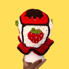 a hand holding up a crocheted hat made to look like a strawberry on top