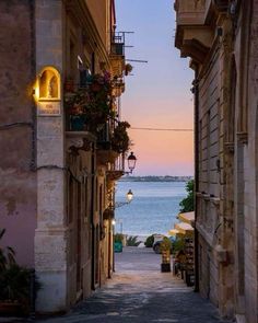 an alley way leading to the ocean and buildings with flowers on them at sunset or dawn