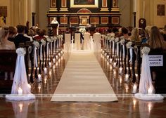 a bride and groom standing at the end of a church aisle with candles in front of them