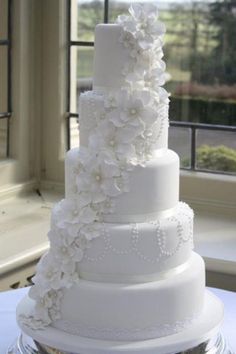 a large white wedding cake with flowers on the top and bottom tier is sitting in front of a window