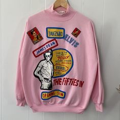 What an amazing piece of fashion history! Made in Canada by Weekend International, this baby pink sweatshirt features a Mockneck and bold artwork. The front is decorated with James Dean printed graphics. Both front and back have Elvis-related decorations which were stitched on by the original owner to make this a fun custom piece. There is some minor wear, including a mark on the yellow front circle graphic (right beside James Dean's head), the odd faint mark, and some fuzzing, but this sweatshi Retro Long Sleeve Tops With Character Print, Retro Long Sleeve Sweatshirt With Cartoon Print, Pink Sweatshirt With Screen Print For Streetwear, Retro Pink Sweatshirt With Graphic Print, Retro Pink Graphic Print Sweatshirt, Pink Graphic Print Sweater For Streetwear, Oversized Pink Top With Cartoon Print, Pink Long Sleeve Sweatshirt With Graphic Print, Pink Graphic Print Sweater For Fall