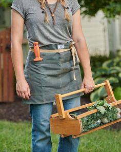 a woman in an apron holding a wooden basket with vegetables inside it and standing on the grass