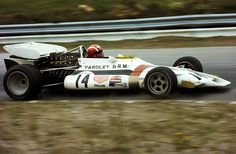 a man driving a race car on a track with grass and bushes in the background