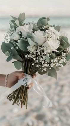 a woman holding a bouquet of white roses and greenery in her hand on the beach