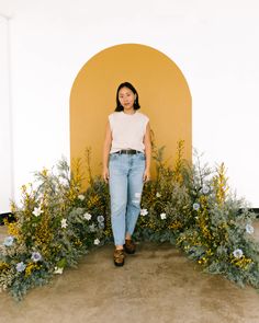 a woman standing in front of a yellow wall with flowers and plants growing around her