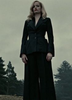 a woman standing on top of a hill wearing a black suit and tie with trees in the background