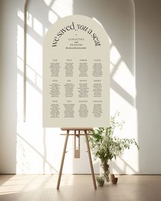 a wedding seating chart on an easel in front of a window with potted plants