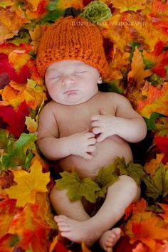 a baby wearing a knitted orange hat laying on top of leaves in the fall