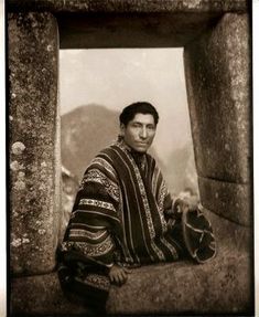 a black and white photo of a man sitting in front of a stone structure with mountains in the background