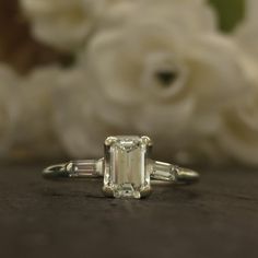 an emerald cut diamond ring sitting on top of a table with white flowers in the background