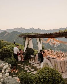 a couple sitting at a table in the middle of an outdoor area with flowers and greenery