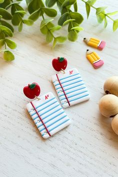 two earrings with pencils and an apple on them sitting next to some green leaves