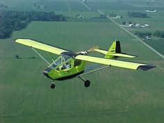 a small yellow airplane flying over a lush green field next to a rural area in the distance