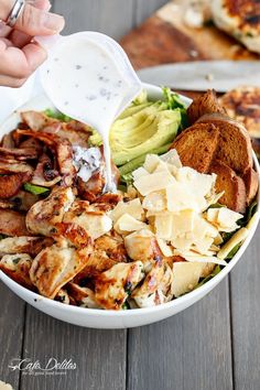 a white bowl filled with chicken, avocado and other food items on top of a wooden table