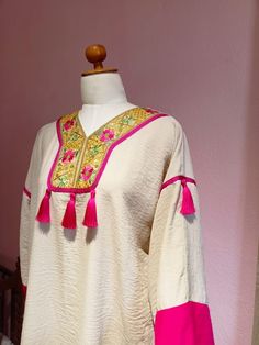 Embrace the charm of traditional Tunisian attire with our "Mony Kamraya" Jebba, a stunning blend of cultural heritage and modern flair. Crafted in a soft, textured fabric, this elegant piece features a rich cream base highlighted with vibrant pink accents. The intricate, hand-embroidered yoke showcases a lively floral pattern adorned with gold thread, while playful tassels add a touch of whimsy. Designed in a one-size-fits-all standard size, this Jebba is perfect for any occasion, ensuring you s Festive Kurta With Tassels, Traditional Pink Kaftan With Traditional Drape, Festive Kurta With Tassels And Traditional Drape, Festive Bollywood Kurta With Tassels, Festive Cream Kaftan With Resham Embroidery, Bollywood Style Kurta With Tassels And Traditional Drape, Bollywood Style Festive Kurta With Tassels, Traditional Kaftan With Tassels, Festival Kurta With Tassels In Traditional Drape