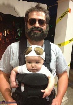 a man is holding a baby in a black carrier and wearing sunglasses on his head